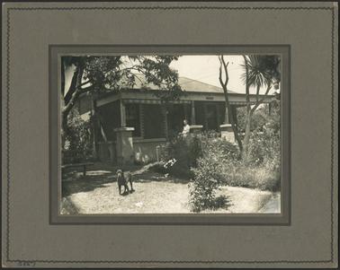 A cement brick home with hipped roof a walled verandah with entranceway into established garden a dog is on the grassed area and a lady is standing on the verandah 