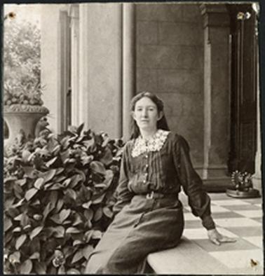 The image is of Leila Steedman sitting on the patterned paving of a verandah, to the left of the photo shrubbery and an urn