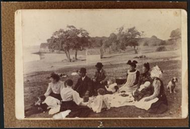 The Sherlock family, which in photo, comprised of seven adults and four children, at a beach at Mornington having a picnic all appear to be looking out to sea, perhaps also a family dog which is also in photo, and in the background are trees and cliffs