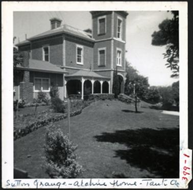 A side view of Sutton Grange c1870s, the house is two storey with a one level concave roof, verandah abutting the square tower the main roof is hipped, established gardens in the foreground