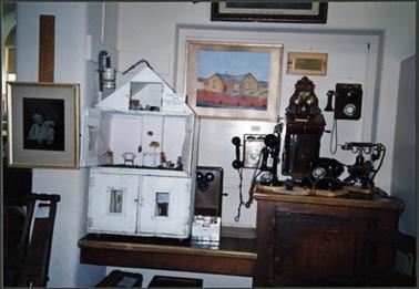 A front view showing Dolls House made from gin boxes 1892, at right side of photo are the Museums collection of antique telephones on wall behind is painting of Museum