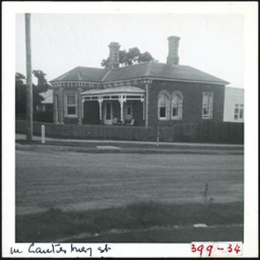 A brick house with a verandah over the porch two windows are arched but all the windows have patterned brickwork around them two chimneys also with patterned brickwork and a wooden fence in front of house