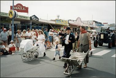 Participants in Australia Day parade 1995 including Neil Taylor and Tessa in period costume, walking past ‘The Mall’ in Main Street, Tessa is pushing pram, and gold miner pushing a wooden wheel barrow. Theme for 1995 was “Transport”