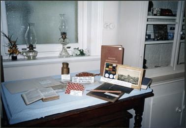 School requisites, displayed on table, on window sill behind table are two kerosene lamps and to right of table is a display cabinet