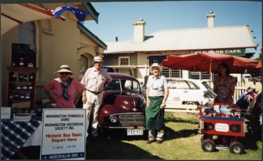 Members of the Mornington Historical Society standing outside the Museum on Australia Day 2002, to the left is M. Platt behind the OPOM stall, then B. Coventry and N. Taylor with the morris car and M. Tregonning with the carousel