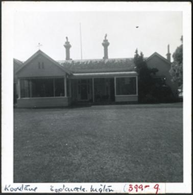The image is of a house with hipped roofs at either end and a flat roof in the centre of the building the 
entrance is in the centre of the building three chimneys can be seen it appears a cross is on the top of the left hipped roof