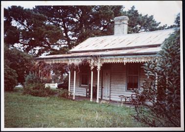 A white wooden cottage with windows on either side of doorway hipped roof one chimney lacework along verandah