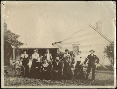 The White family, being seven females and two males, photographed in front of a hipped roof house which has a verandah to the left of the building, female standing at the end (left) is holding onto a bicycle and a male standing at end (right) also holding onto a bicycle