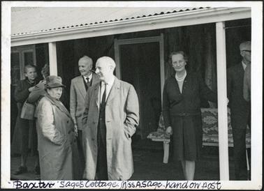 The image is of a group of people standing under the verandah at Sages Cottage, this includes Miss Annie Sage (with her hand on verandah post)