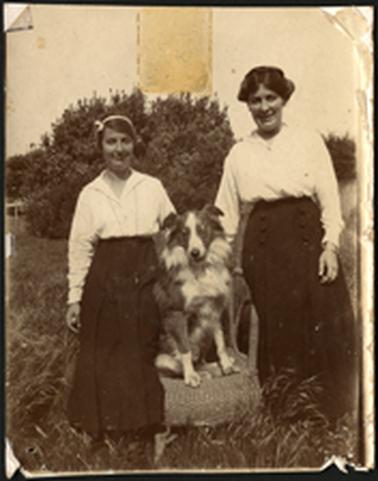 The image is of Dora and Rene Bates in an outside setting, standing on either side of their dog which is seated on a cane chair