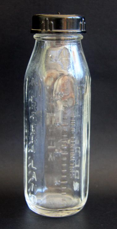 Glass Pyrex baby’s bottle with bakelite screw top lid. Teat held inverted to be ready to be attached for feeding. 