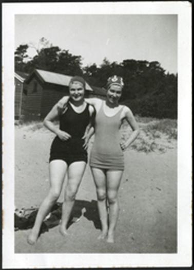 Two females (Stone?) in bathing costumes standing on a beach with a bathing box to the left of photo behind them