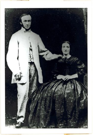 This shows a couple in Victorian dress. The gentleman is standing and the woman is sitting down. The gentleman is wearing a light coloured long jacket with a shirt with a stand up collar, and a bow tie. The woman is wearing a dark dress with a white lace collar. 