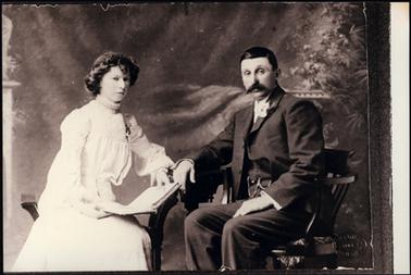 This photograph has been taken in a studio, and shows a man and woman seated. The man is on the right of the picture and he is very smartly dressed in a dark three piece suit, with a watch chain and flower button hole. The woman is in a white dress, with smocking to the high neck, and the top of each sleeve. She is holding an open book. Her hair has been carefully curled.