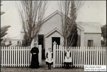 This photograph shows a white one storey building, with a pitched roof. The rear section of the building is weather board, but the front porch appears to be rendered. There is a white picket fence at the front with a gate way. There are two tall trees either side of the gate on the inside. In front of the fence there is a  woman and two small girls. The woman teacher is on the left, and is wearing a dark long dress. The two pupils are wearing dark dresses with white pinafores over them. They have dark stockings and shoes.
