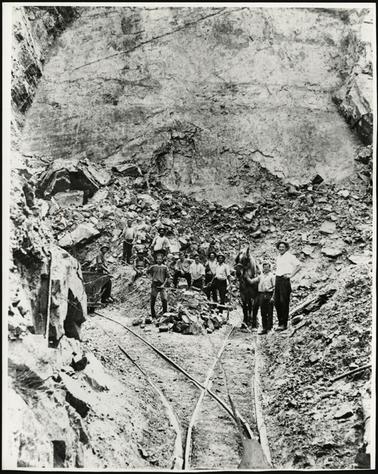 This image shows a group of workers at the Quarry face. They are in shirt sleeves, and some are wearing hats. Some of the group are holding tools. There is a horse to the right of the picture, and one of the men is holding him by the bridle. To the right of the photo is a truck which runs on rails or tracks, presumably pulled by the horse when full of rock. 