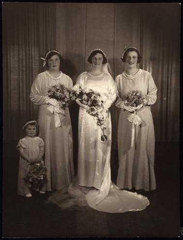 This photograph shows a wedding party of four. The bride, and three bridesmaids. Two of the bridesmaids are adults, and the third a little girl, maybe a maid of honour. Each one has a long gown and the three adults have long sleeves and gloves. They are carrying huge bouquets. The little girl has a long gown with short puff sleeves. The bride has a long train, and this has been spread in the foreground of the picture.