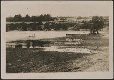 This is a view looking across Mills Beach, with a view of the sea. There is a boat in the centre with two people on board. There is a wooden jetty to the right of the picture.