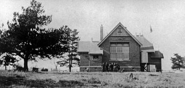 A free standing, single storied brick building on generous sized land. There are at least three rooms off set behind one other.  All have steeply pitched roofs. The style is typical of turn-of-the-century school buildings. Two trees stand to the left and there are six children grouped in the front of the building