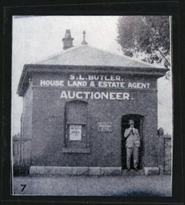 This image shows a small rectangular brick building, with a tiled roof. The base of the building appears to be blue stone blocks. The doorway is to the right, the one sash window is to the left. One chimney can be seen. There is a gentleman stood in the doorway wearing a suit and tie, and a Trilby hat. There is a sign painted on to the brick above the door and window “S.L.Butler, House, Land & Estate Agent. Auctioneer”.