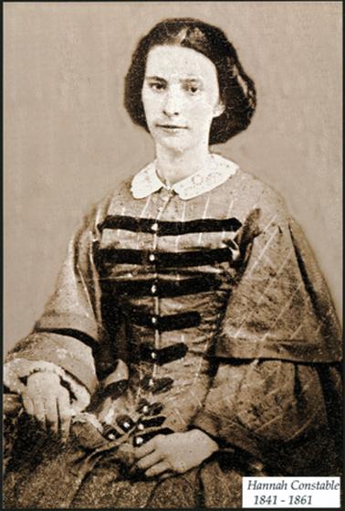 This image shows a woman with black hair with a centre parting. She is in period dress. Her dress has a lace collar, and black strips across the bodice where the buttons are. This is a studio photo.