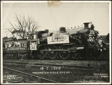 The Mornington Train decorated for 'Peace' and Victory', a sign on top of the train reads 'Rejoicing From Mornington' and another sign behind the driver's compartment reads 'Our Allies Rejoice With Us', flowers and flags also adorn the train