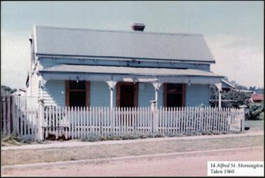 This photograph shows a white weather board house. It is single storey with a pitched corrugated iron roof. There is a verandah across the front of the property, with a ornamental trim. There is a central door with a sash window either side. the door and window frames are a timber colour. There is a picket fence to the front of the property.