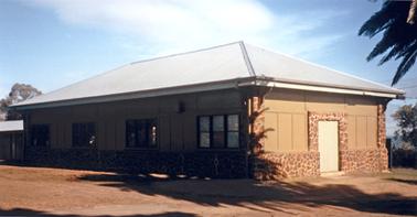 A rectangular shaped building which has been constructed of red stone around the base and lathe and plaster exterior walls painted in an ochre colour. The roof is a simple skillion shape made from corrugated iron. There is a door at one end and a series of windows and doors along the front face. There is a shadow of palm tree fronds in the upper right hand corner.