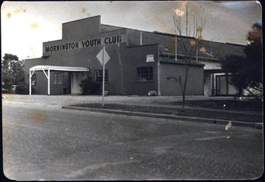 This photograph shows a large brick fronted building which has a large sign “Mornington Youth “Club” in black lettering on the front. It has a portico painted white over the front entrance. On either side of the entrance are two windows each side. The roof is corrugated iron.