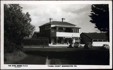 This is a photograph of a Rose Series post card showing an impressive two storey brick and stone hotel. The name of the hotel is on the side of the building and above the entry. There is a verandah to both levels, this is made of wood. The top floor verandah is partially enclosed. The roof is slate, and two chimneys are visible.There is a car parked outside, and people sitting outside on the ground floor level.