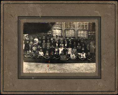 This photo shows 56 pupils and 2 teachers, one on either side. There are in 4 graduated rows, with the children in the front row cross legged. The little boy in the centre front is holding a sign “Mornington School 2033 Dec. 1938, Grades 1 & 2. 