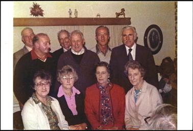 This shows a total of ten people, six men standing and four women sitting. They are inside a house. On the wall to the right of the group is an oval frame showing a couple. There is also a woman sitting aside, and hiding her face with a hat.