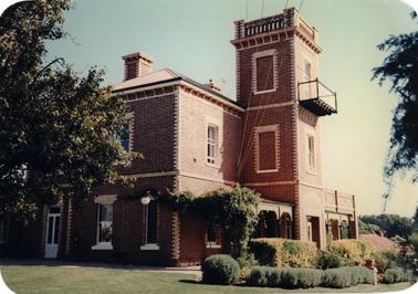 This is a photograph of a distinguished two story brick house with a three story tower against the front wall. Iron lace frames a verandah on either side of the tower which has a lookout balcony on its third story. The house is set in a formal garden with a lawn and low hedges. There is an entrance in the side wall as well as the front entrance through the ground floor of the tower. Tall windows are framed in decorative tuck pointed brick.  