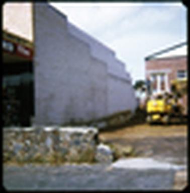 This photograph shows the side of a shop. It is a tall brick wall, painted white. In the foreground is a small rock wall. In the background, there is a glimpse of a yellow painted industrial truck of some sort, with caterpillar tyres. There is also a man stood watching.