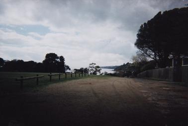 This photo shows an unmade road leading down to the beach in the vicinity of St McCartans. It has a green playing field on the left with a pine fence. On the right there is a paling fence and a gate. 