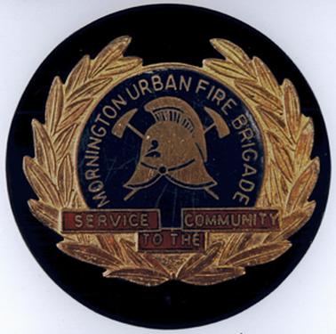 A circular badge has a black background containing a golden laurel wreath with a fireman’s helmet placed centrally surrounded by “MORNINGTON URBAN FIRE BRIGADE” and “SERVICE TO THE COMMUNITY “below.