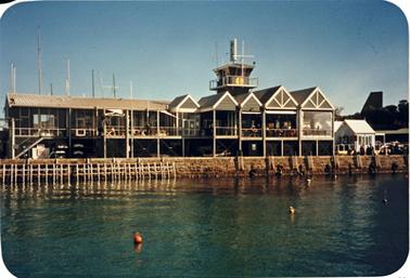 This image shows a view from the water of the Yacht Club. It has an interesting roof line, with five pitched sections. There is a tower behind. It appears to be of timber construction and two storey. To the right of the photo there is a separate small building with a pitched roof. Over the roof of the club the masts of the yachts can be seen.