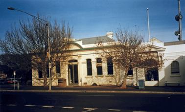 A  building from the late 19th century, now modernised, stands on a street corner. Its original walls are now rendered white. The main building is symmetrical about a front entrance, with three windows either side of a main door. The door frame is in modern style. An adjoining building has a lower roof line, one window on either side of a former entrance and is similarly rendered. The word MEMORIAL in raised lettering is on the wall above the window on the left. There are two deciduous trees in the street and the street furniture includes a seat, a memorial bollard and a decorative container for garbage. 
