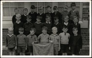 This shows 22 young boys arranged in 3 tiers. The boy in the centre of the front row is holding a banner with the number 2033 embroidered in the centre. The grade number is not visible. Some of the boys are in uniform, and some are not. The boys are stood in front of the school building.