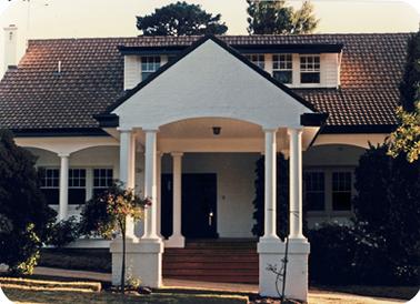 A double fronted white house with a trio of casement windows on each side of the main entrance and a second floor room with similar windows  is set in the tiled roof. The front door opens from a verandah which is defined by slim, white pillars and approached from the driveway by a set of terracotta steps. The driveway in front is spanned by a portico of similar white pillars.