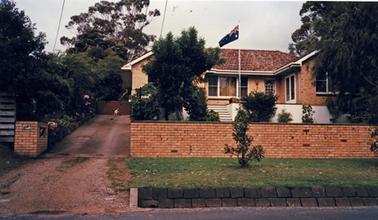 A single storied cream brick house stands in a small garden behind a low solid brick fence with an open driveway of gravel beside the house. It is an L-shape with a set of steps up to a terrace defined by a low white stucco wall. There are several windows, a single door and a double French window on the front facade. A flag pole flying the Australian flag stands in front of the terrace. A small dog is standing at the end of the drive which is lined by a garden strip along the side fence. There are several shrubs and trees in the garden and a grassed nature strip containing a young shrub.