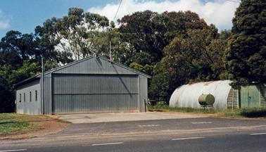 Two sheds stand together on flat land beside a road and in front of a stand of mature trees. The shed on the left is a large, grey square building with a gabled roof. It’s facade is grey vertical corrugated iron surrounding a very wide door. The shed on the right is the old Nissen hut which was the first Moorooduc Fire Station (see 1288.1). 