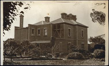 This photograph shows a grand two storey brick building from the rear. It has a tower which can be seen over the roof top. The roof is slate. The sash windows are surrounded with a decorative brick effect in white which contrasts with the surrounding brick. 
