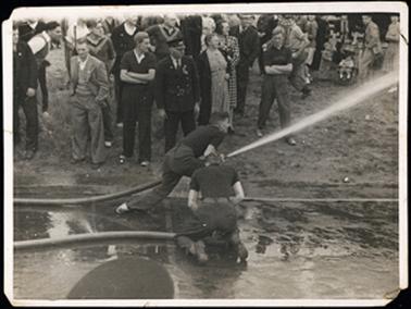 This photograph shows two men demonstrating the use of the big hoses used to fight fires. One hose is actually shooting water. There are a crowd of onlookers, mostly looking in the direction the hose is pointing. 