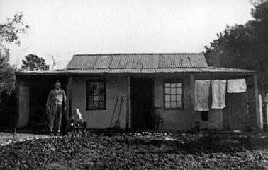 Rudimentary wattle and daub house with corrugated iron roof and 2 multi - paned windows either side of a central door. There is a man in work clothes standing beside a white cockatoo on the left and some clothes hanging on a line under the verandah on the right. 