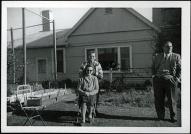 This image shows two women and a man in the garden at Tasma Court. One woman is seated and the other is standing behind her chair. The gentleman is standing apart to the very right of the picture. He is wearing a three piece suite, shirt and tie. They all have spectacles. There is a building directly behind them, which looks to be built of weather board and fibro sheeting.
