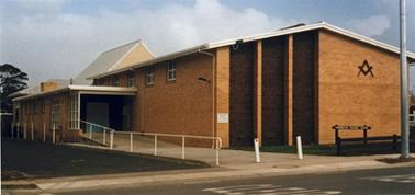 A substantial cream brick rectangular building has a low gabled roof and a small entrance building on the left hand side. There are four small windows high on the side wall and three dark vertical windows occupy the left half of the front facade. The Freemason’s emblem is attached centrally in the upper part of the right half. A wide paved access path leads up to the doorway from the road in front. Two white posts near the public footpath limit vehicle access and there is a white hand rail beside the path to the main entrance. Attached to the rear of the main hall is a small high gabled building and the lower roof of another. A simple brown wooden sign in front identifies the ‘Mornington Masonic Centre’.