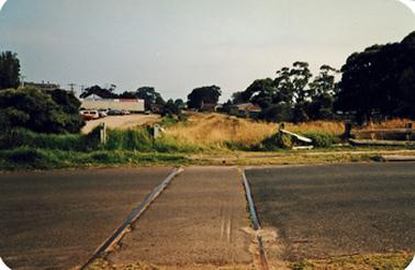 A view across vacant land with a roadway in the foreground and a pair of railway lines across it. In the background are commercial buildings and parked vehicles on the left and a few non-specific buildings  among a line of trees on the right.