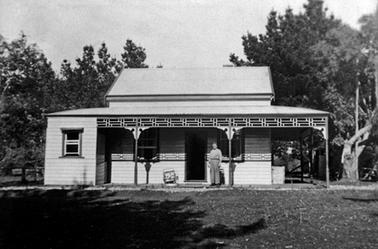 Painted weather board house with a window on each side of the main door. On the left is a small enclosed sleepout with a door on to the front verandah. It has a skillion roof of corrugated iron.