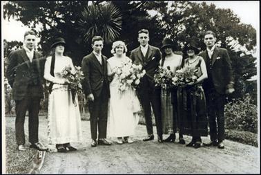 This photograph shows a wedding party in the driveway at Wolfdene. It consists of the bride and groom and 3 bridesmaids, and 3 groomsmen. The women are carrying large bouquets. The bride has an ankle length long white gown, and each of the bridesmaids appears to be wearing ankle length gowns of different colours. The bridesmaids are wearing cloche hats, and the bride a circlet of white flowers. The men are wearing dark 3 piece suits with white stand up collars and white ties.